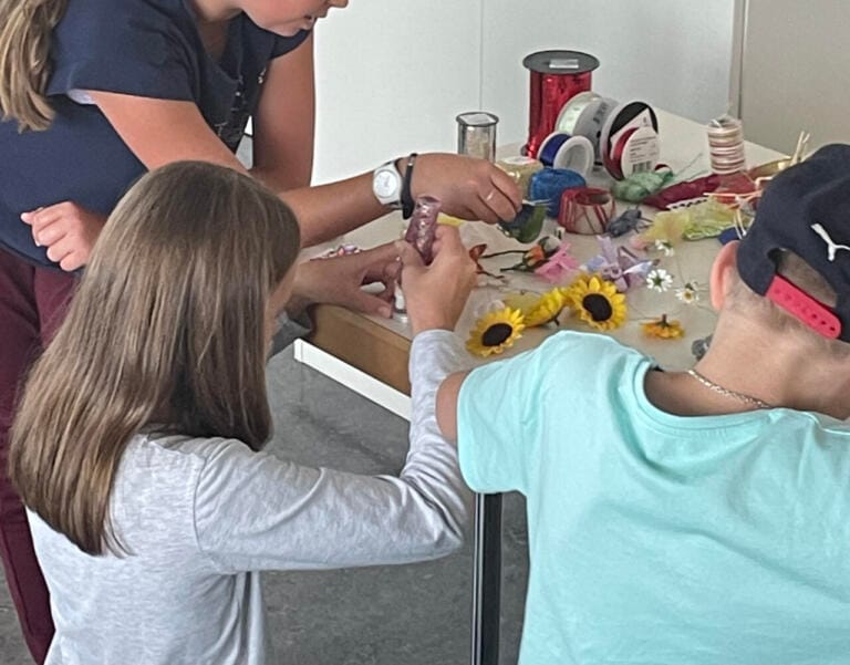 Drei Kinder sitzen und stehen um einen Tisch und basteln mit künstlichen Sonnenblumen, bunten Bändern, Garn und anderen kreativen Materialien. Ihre Gesichter sind nicht zu sehen. Die unordentliche Oberfläche ist übersät mit bunten Bastelutensilien.
