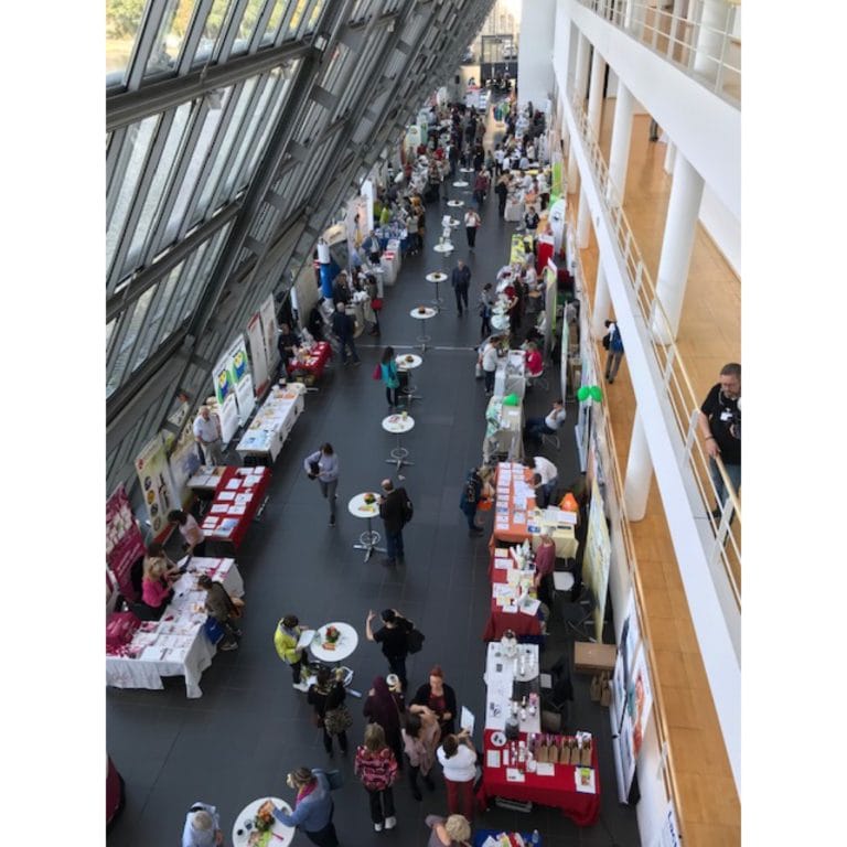 Luftaufnahme einer geschäftigen Hallenausstellung oder Messe in Gelsenkirchen, mit Menschen, die zwischen Ausstellungstischen und Ständen in einer großen, sonnendurchfluteten Halle mit Glaswänden und hohen Decken umhergehen.