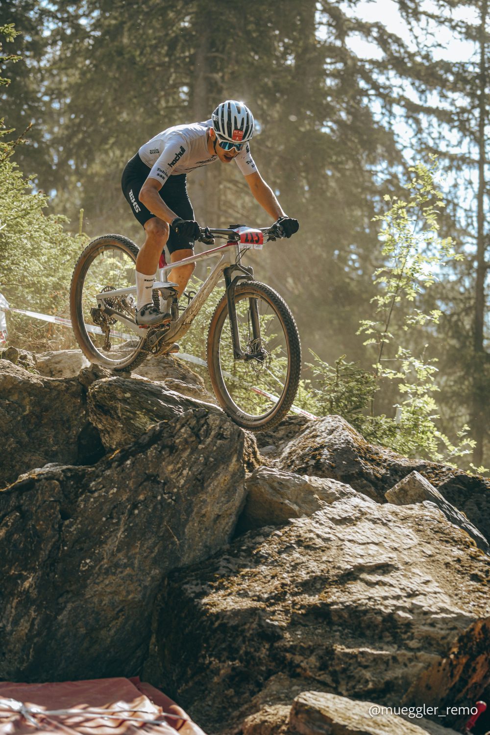 Ein Radfahrer mit Helm und Rennausrüstung fährt mit einem E-Bike einen steinigen Waldweg hinunter und umfährt im hellen Sonnenlicht geschickt große Felsbrocken.