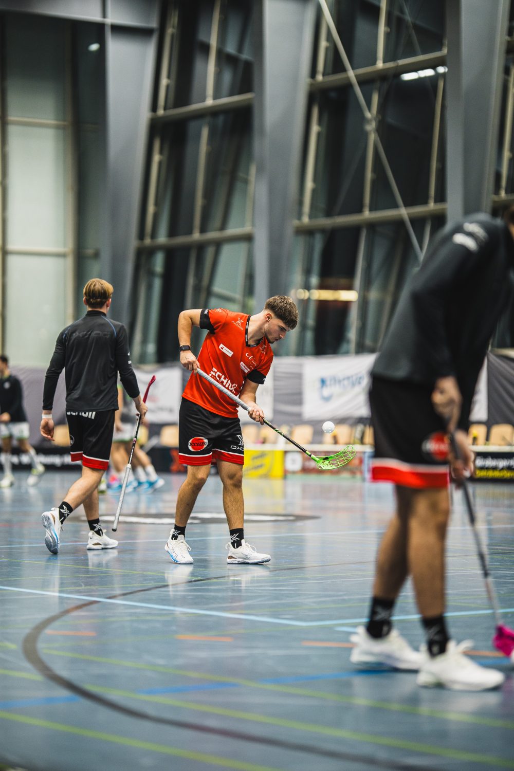 Drei männliche Unihockeyspieler in Sportuniformen, darunter Josia Pfister, stehen mit Unihockeyschlägern in der Hand auf einem Hallenplatz. Der Hauptspieler im roten Trikot wirkt konzentriert und einsatzbereit, im Hintergrund sind große Fenster und Arenalichter zu sehen.