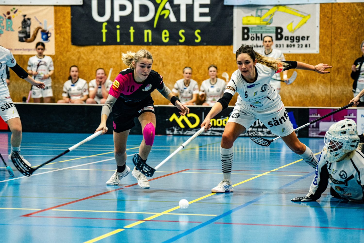 Leonie Soraya Mai stürzt sich nach vorne, während zwei Unihockeyspielerinnen in einer Halle intensiv um den Ball kämpfen. Im Hintergrund sind weitere Spielerinnen und ein Torwart zu sehen, die die sportliche Szene ergänzen.