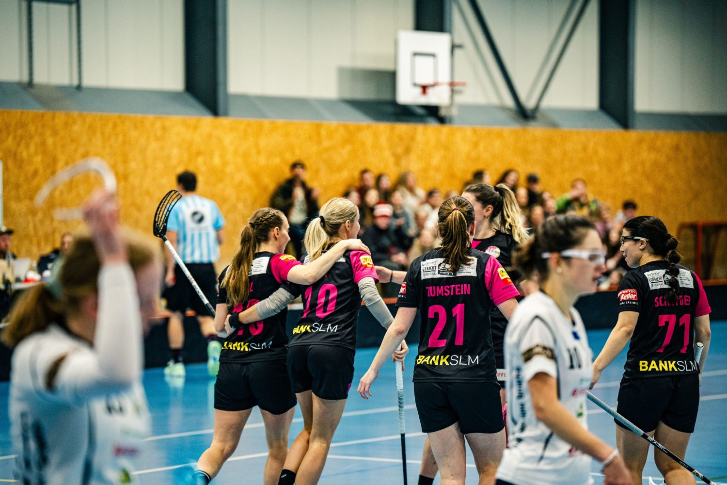 Ein Frauen-Unihockeyteam in schwarzen Trikots feiert auf einem Hallenplatz, die Arme umeinander gelegt, während Spielerinnen in Weiß daneben laufen. Die Zuschauer beobachten, wie Leonie Soraya Mai und ihre Mannschaftskameradinnen den Moment teilen.