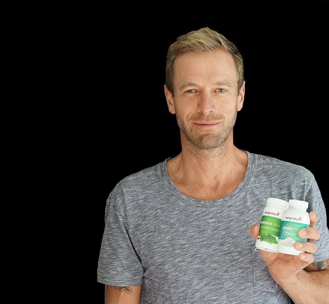 Matthias Larsen, ein Mann mit kurzen blonden Haaren und einem grauen T-Shirt, hält vor einem schlichten schwarzen Hintergrund zwei Flaschen mit Nahrungsergänzungsmitteln hoch.