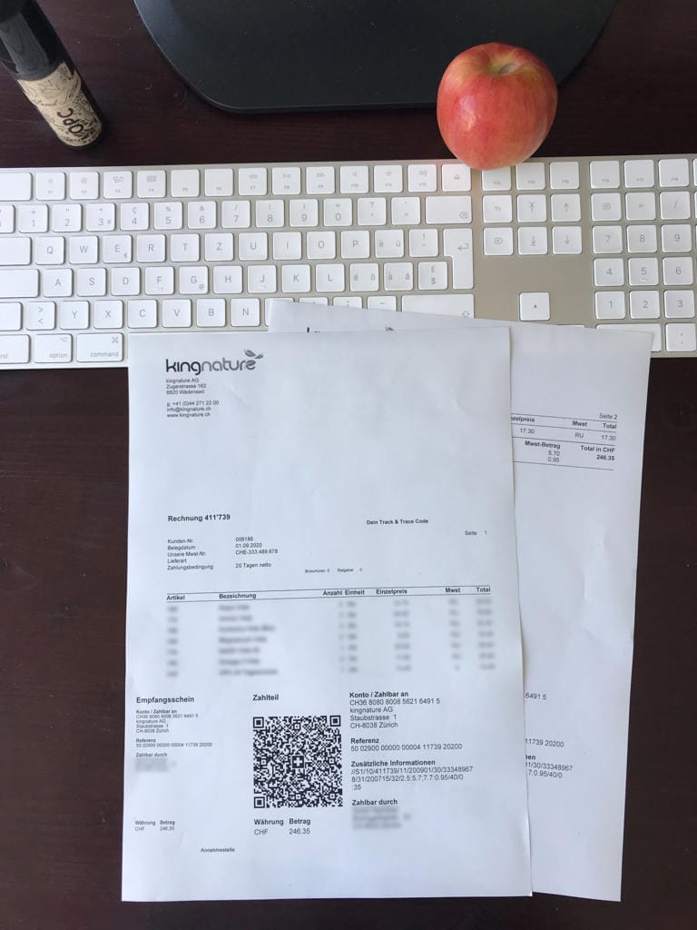 Zwei Rechnungen von Kingnature mit QR-Codes liegen auf einem Schreibtisch mit weißer Tastatur, schwarzem Monitorständer, Apfel und kleiner Flasche. Die mit „Rechnung mit QR-Code“ beschrifteten Papiere verdecken teilweise die Tastatur.