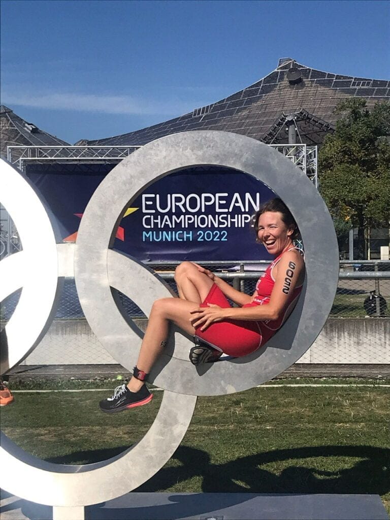Eine Sportlerin im roten Triathlonanzug posiert lächelnd in einer großen Skulptur der Olympischen Ringe. Hinter ihr steht ein Schild mit der Aufschrift „Europameisterschaften München 2022“, im Hintergrund das markante Gebäude mit Glasdach.