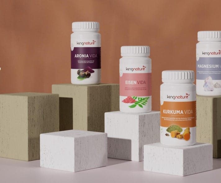 Drei Flaschen Nahrungsergänzungsmittel von kingnature AG – Aronia Vida, Eisen Vida und Kurkuma Vida – werden auf Holzklötzen vor einem braunen Hintergrund präsentiert, wobei rechts ein Teil einer vierten Flasche mit der Aufschrift „Magnesium“ zu sehen ist.
