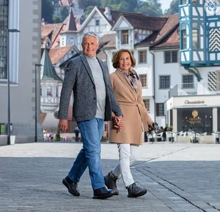 Ein älteres Paar hält Händchen und lächelt, während es auf einer Kopfsteinpflasterstraße in Zürich spazieren geht und im Hintergrund die historischen Gebäude dieser malerischen europäischen Stadt genießt.