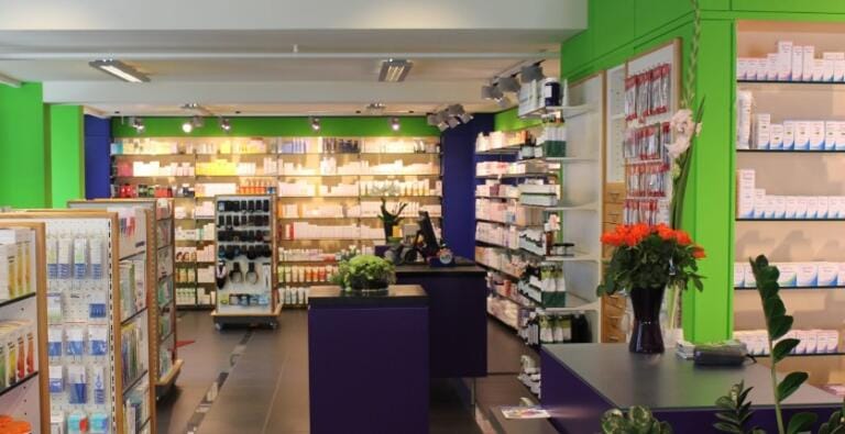 Ein helles Interieur der Dorfdrogerie Baumann mit grünen und violetten Wänden, Regalen voller Gesundheitsprodukte, einer Theke mit einer Vase mit orangefarbenen Blumen und geordneten Auslagen im gesamten gut beleuchteten Raum.