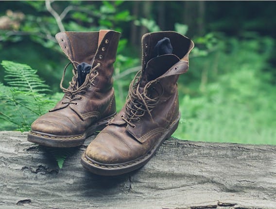 Ein Paar abgetragene braune Lederstiefel mit offenen Schnürsenkeln stehen auf einem umgestürzten Baumstamm in einem üppigen grünen Wald und rufen das Gefühl von Naturfuß und Fürsorge hervor, das man in einer Fußpflegepraxis findet, umgeben von Farnen und verschwommenem Laub im Hintergrund.