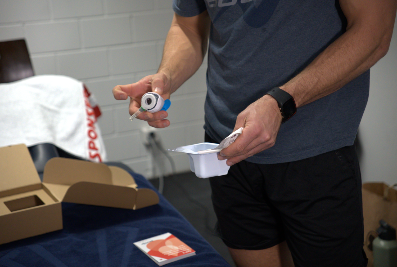 Eine Person in Sportkleidung, möglicherweise ein Schweizer Eishockeyfan, hält einen Metallkanister und einen weißen Kunststoffbehälter mit abziehbarem Deckel, der neben einer offenen Schachtel auf einer blauen Fläche steht. Im Hintergrund sind ein Handtuch und verschiedene Gegenstände zu sehen.