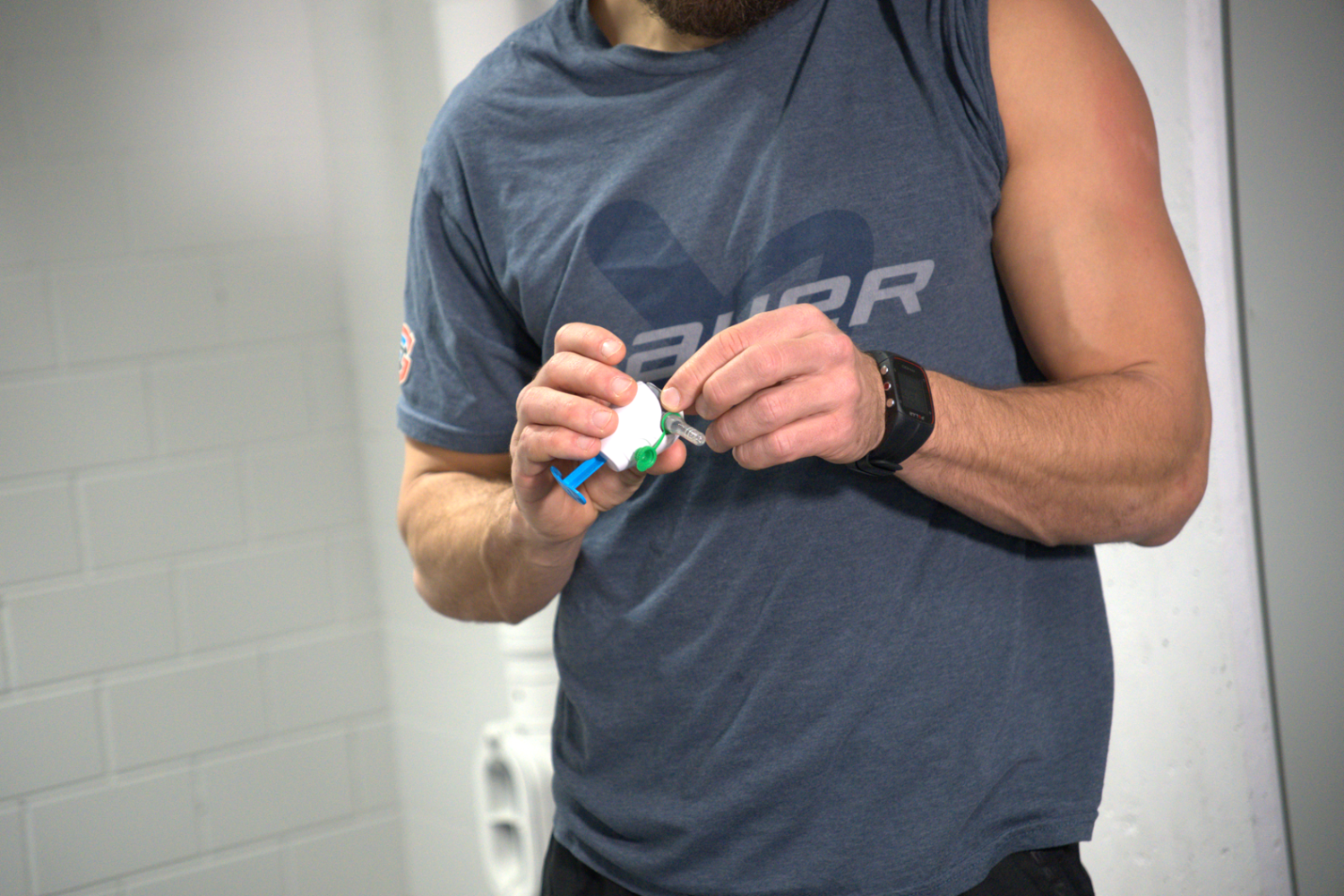 Eine Person in einem blauen ärmellosen Hemd und einer schwarzen Uhr hält einen weißen Inhalator in der Hand und macht sich bereit, ihn zu benutzen. Der Hintergrund, der an eine Schweizer Eishockey-Arena erinnert, zeigt eine weiße Wand mit Ziegeln und einigen Sanitäreinrichtungen.