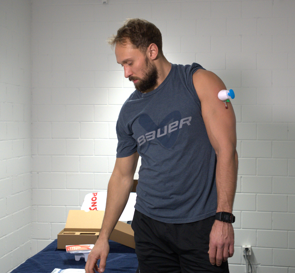 Ein Mann in einem blauen HC Fribourg-Gottéron-T-Shirt steht in einem Haus mit einem medizinischen Gerät am Oberarm und blickt auf die Materialien auf einem Tisch. Im Hintergrund sind eine weiße Backsteinmauer und Pappkartons zu sehen.