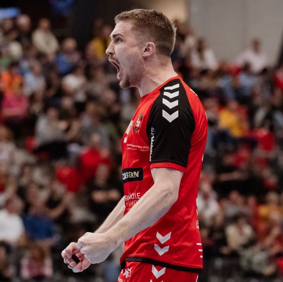 Simon Getzmann, ein männlicher Athlet in rot-schwarzer Uniform, feiert leidenschaftlich – er ballt die Fäuste und schreit. Eine verschwommene Menschenmenge füllt den Hintergrund und unterstreicht die Intensität des Sportereignisses.