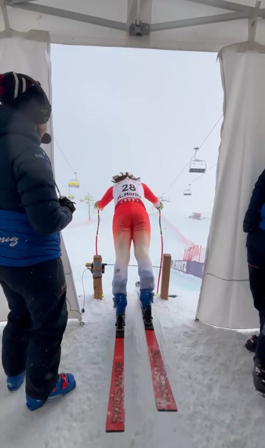 Allegra Frei, in rot-weißer Kleidung mit der Startnummer 28, bereitet sich auf den Start eines Abfahrtsrennens vor, während zwei Offizielle in der Nähe stehen und die Skilifte im Hintergrund zu sehen sind.