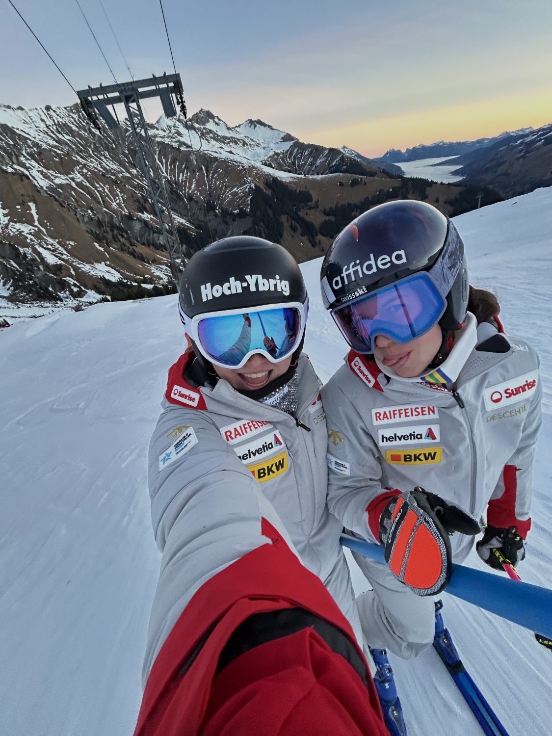 Zwei Skifahrer in weissen Anzügen und Helmen, darunter Allegra Frei, machen ein Selfie auf einer schneebedeckten Bergpiste bei Sonnenuntergang, mit Skiliften, Bergen und einem bunten Himmel im Hintergrund.