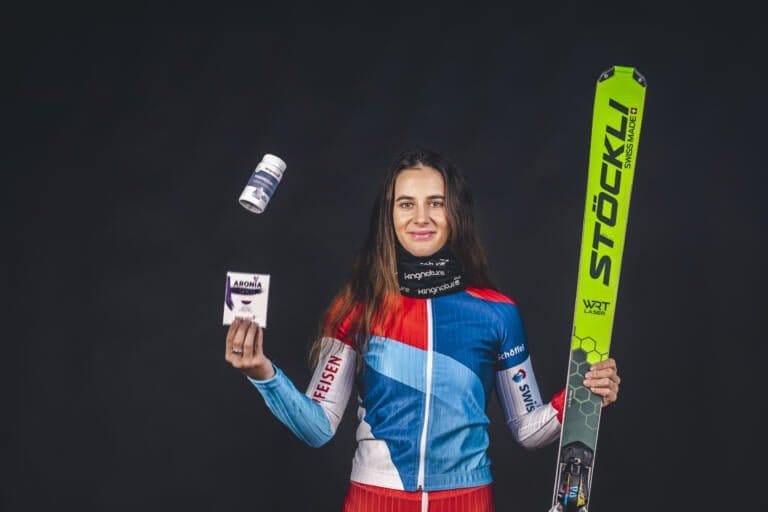 Talina Gantenbein, eine Skicrosserin im bunten Skianzug, hält in einer Hand einen grünen Ski, während sie mit der anderen Hand einen Behälter, eine Flasche und eine Schachtel jongliert und vor einem dunklen Hintergrund lächelt.