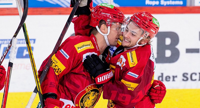Two ice hockey players in red uniforms, helmets, and gloves celebrate on the ice, smiling and embracing each other after a play. The background shows advertisements on the rink boards.