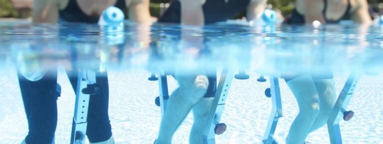 Mehrere Personen nehmen an einem Aquafitnesskurs teil und fahren auf Heimtrainern, die teilweise in einem Schwimmbecken untergetaucht sind. Die Wasserlinie ist im gesamten Bild sichtbar.