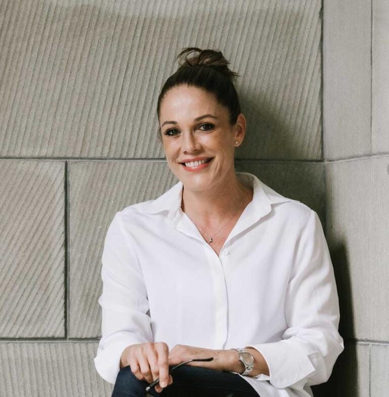 Eine Frau mit dunklem Haar zu einem Dutt, einem weißen Hemd mit Knöpfen und einer Uhr sitzt lächelnd vor dem Hintergrund einer Steinmauer und strahlt damit Vertrauen in Coaching und Psychologie aus.