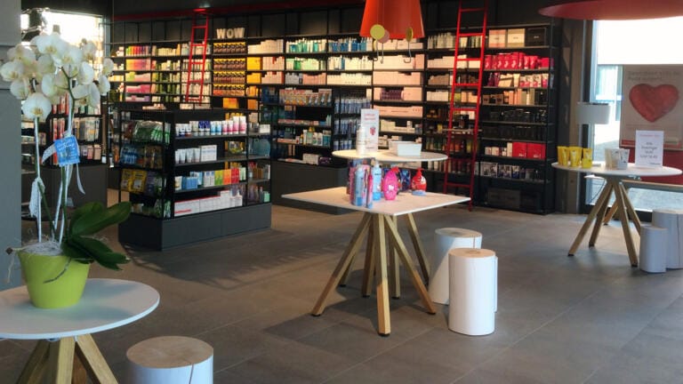 Ein helles, modernes TopPharm-Geschäft mit Regalen voller farbenfroher Schönheits- und Hautpflegeprodukte. Kleine Tische präsentieren Geschenkartikel und Blumen, während rote Leitern und Hängelampen dekorative Akzente setzen.