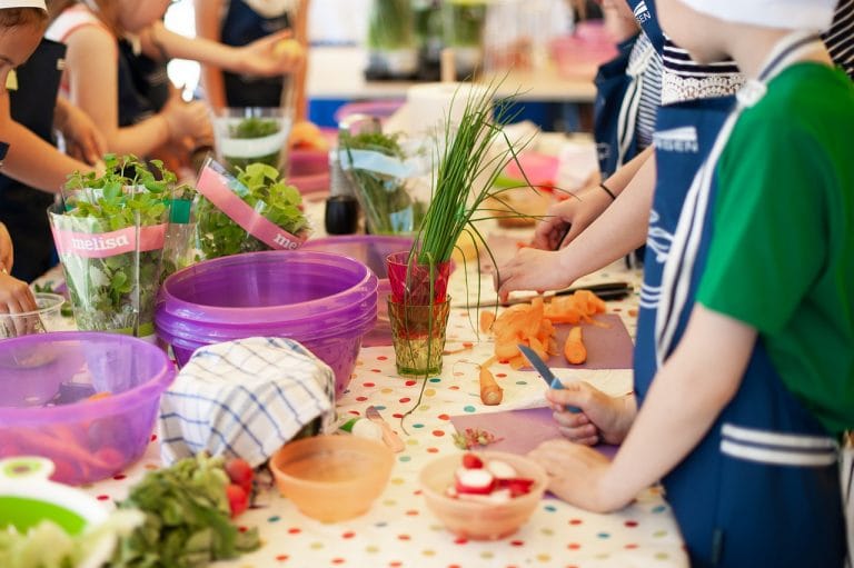 Kinder in Schürzen bereiten gemeinsam Gemüse an einem bunten Tisch voller Schüsseln, frischer Kräuter und Küchenutensilien zu, was eine gemeinsame Kochaktivität nach dem Vorbild der Ernährungsberatung mit Christine Binder nahelegt.