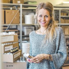 Eine lächelnde Frau mit langen blonden Haaren und einem grauen Pullover steht in einem Lagerraum und hält kleine Flaschen in der Hand. Im Hintergrund sind Kisten und Regale mit verschiedenen Naturprodukten zu sehen.