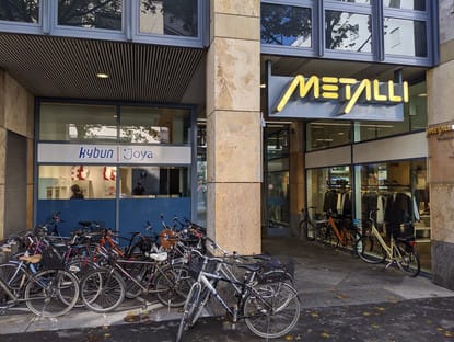Vor einem Gebäude mit großen Fenstern stehen mehrere Fahrräder auf dem Gehsteig. Über den Eingängen hängen Schilder mit den Aufschriften „Joya“ und „Kybun“, im Inneren des Metalli-Ladens sind Kleiderständer zu sehen.
