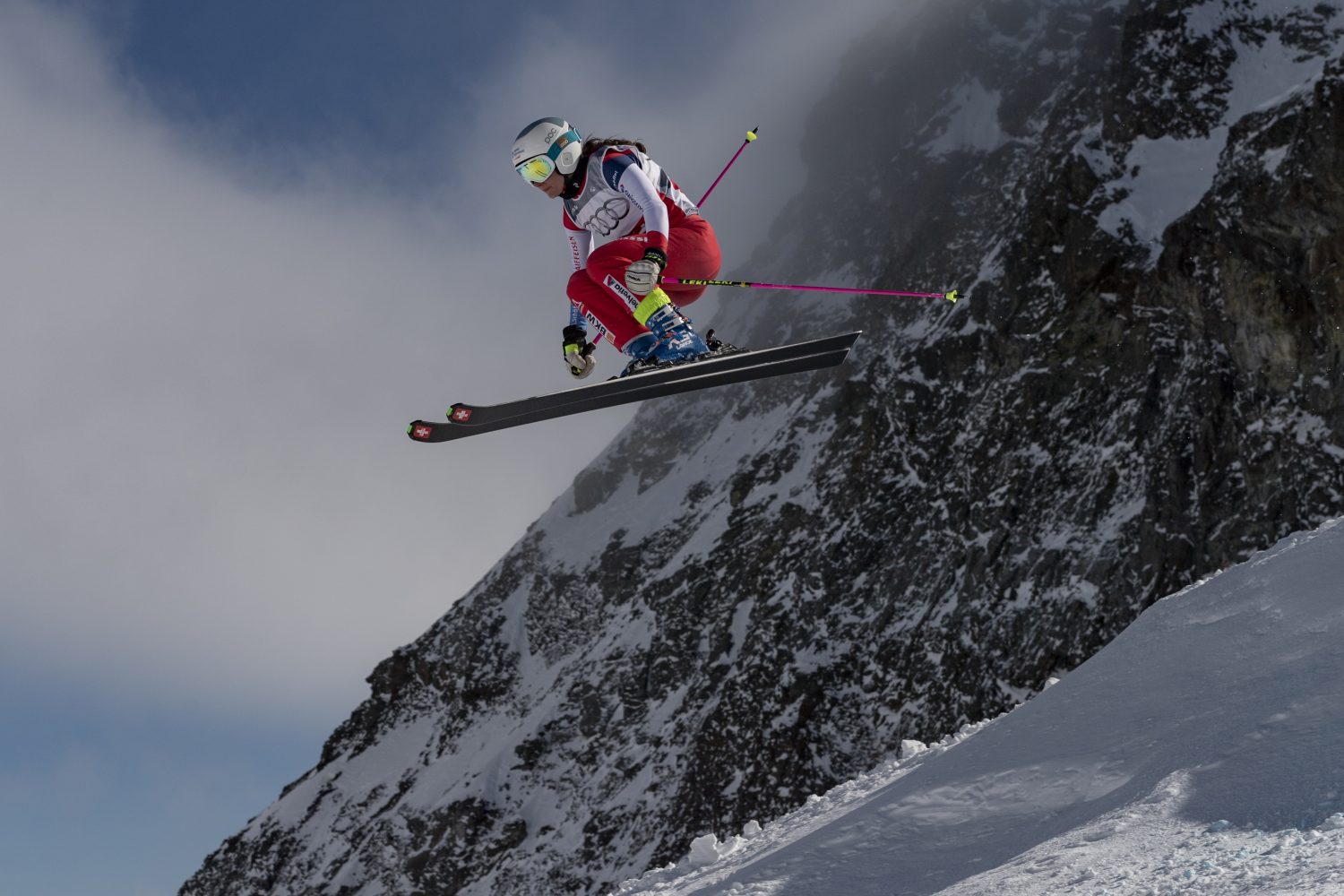 Ein Skicross-Athlet in rot-weißer Ausrüstung schwebt über einem schneebedeckten Berghang durch die Luft, mit nach hinten abgewinkelten Skistöcken und einem felsigen, schneebedeckten Gipfel im Hintergrund.