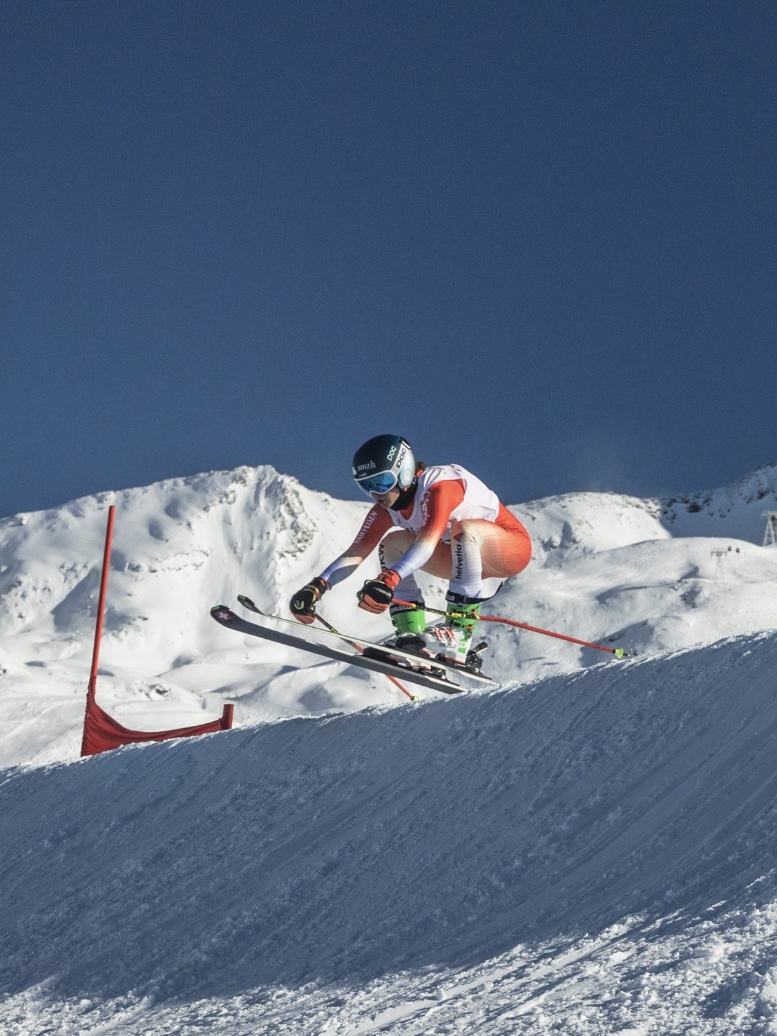 A skier in orange and white gear and a blue helmet races downhill on a snowy mountain slope under a clear blue sky, with red markers in the background.