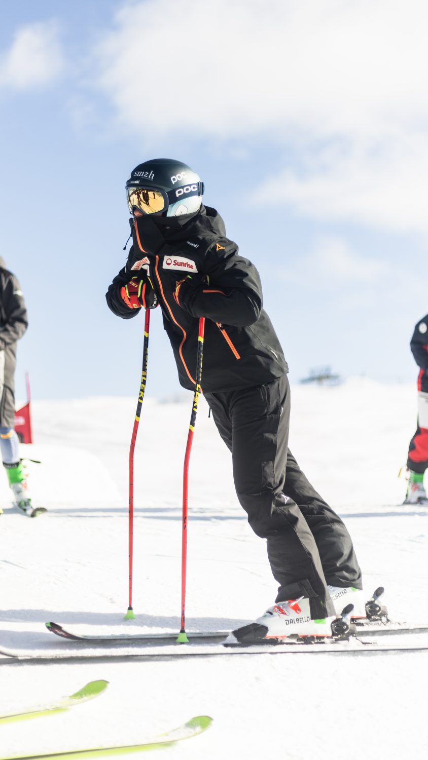 A skier in black gear and a helmet leans forward on their ski poles, preparing to start on a snowy slope under a blue sky. Other skiers are visible in the background.
