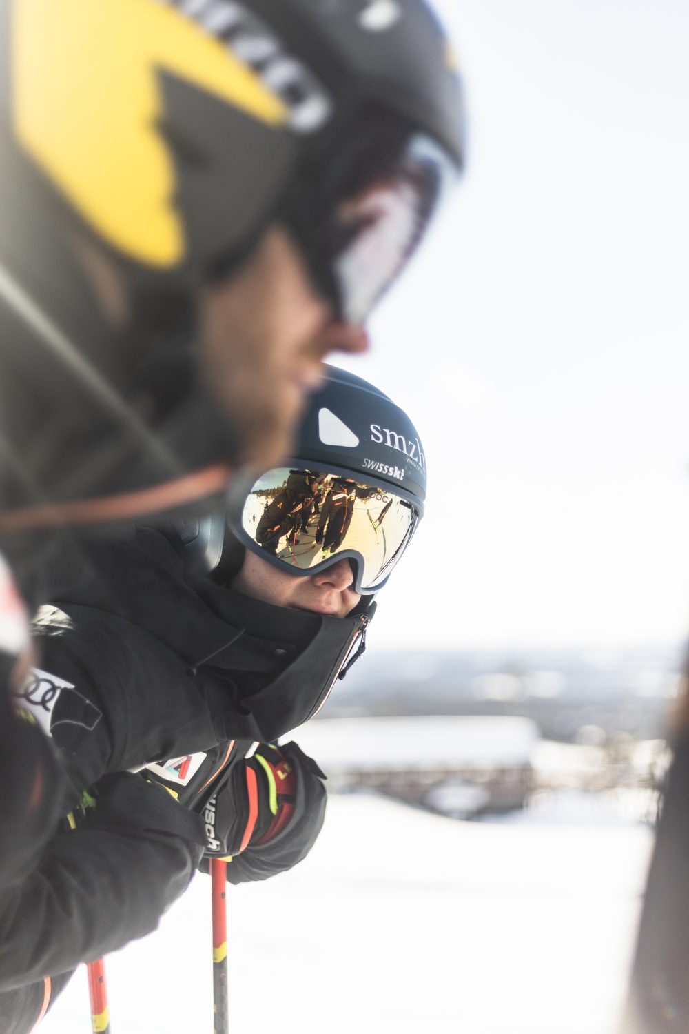 Two skiers in helmets and goggles stand close together on a snowy slope, holding ski poles. The focus is on the skier in the background, while the foreground skier is blurred. Bright, sunny conditions.