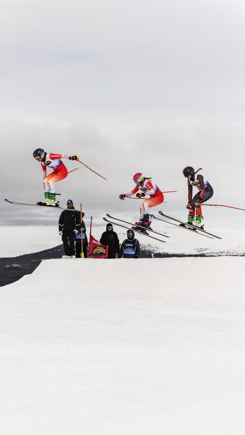 Three skiers wearing helmets and race suits are airborne as they jump off a snowy slope during a ski race, with officials and a course marker visible below against a cloudy sky.