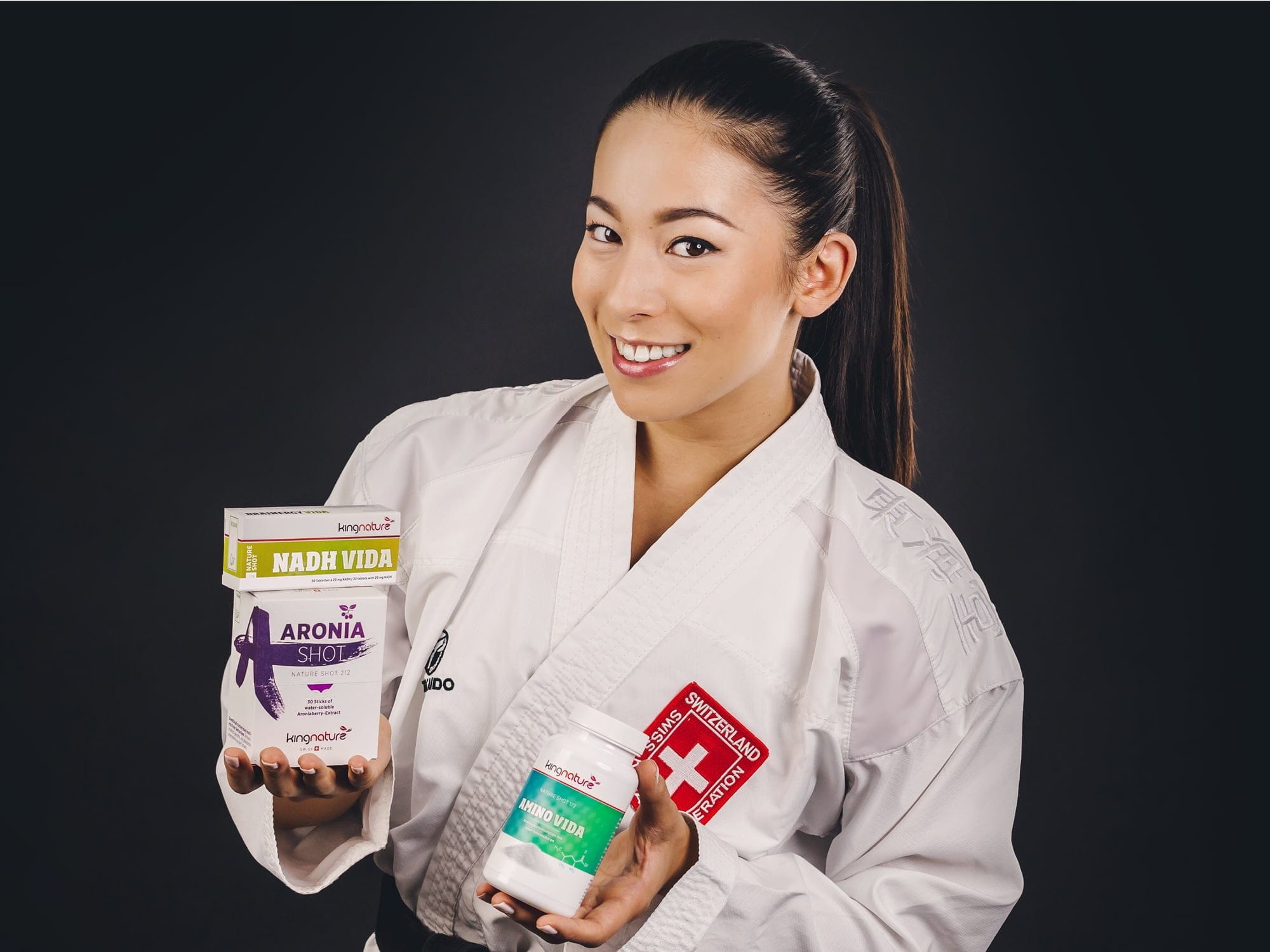 Eine Frau in einer weißen Kampfsportuniform lächelt und präsentiert stolz drei Nahrungsergänzungsmittel vor einem dunklen Hintergrund.