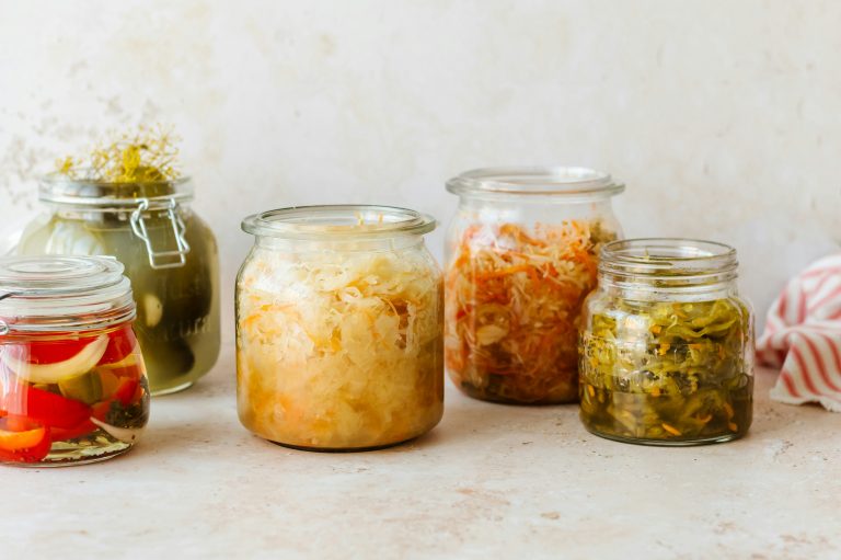 Fünf Gläser gefüllt mit verschiedenen eingelegten und fermentierten Gemüsesorten, darunter Gurken, Kohl und Paprika, heben die Unterschiede zwischen Präbiotika und Probiotika auf einer leicht strukturierten Oberfläche vor einem neutralen Hintergrund hervor.