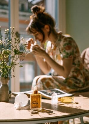 Eine Frau in einem geblümten Kleid sitzt an einem sonnigen Fenster und riecht an etwas in ihren Händen. Auf dem Tisch stehen Drogerie-Parfümflaschen, eine Vase mit Wildblumen und eine Zeitschrift, die die frische Meeresbrise widerspiegelt.