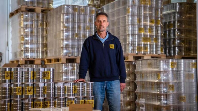 Ein Mann in einem blauen Pullover steht in einem hell erleuchteten, ordentlichen Lagerhaus, umgeben von großen Stapeln von SicherSatt-Metalldosen mit der Aufschrift „SÜSSES SALZ“ auf Holzpaletten.