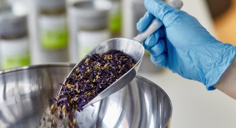 In der Drogerie Schnellmann hält eine behandschuhte Hand eine mit getrockneten violetten und grünen Kräutern gefüllte Metallschaufel über einer großen Edelstahlschüssel. Im unscharfen Hintergrund sind Gläser zu sehen.